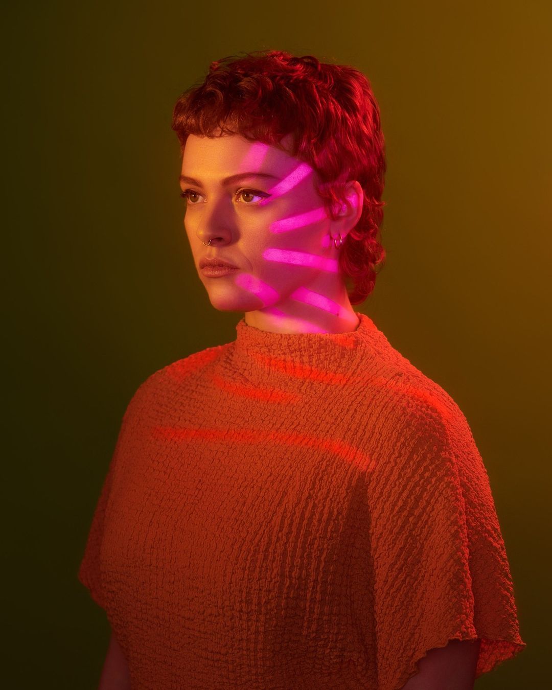 Portrait of a young adult woman with short red hair and a textured shirt, highlighted with pink light stripes against a green backdrop.